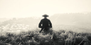 Der Held in Trek to Yomi sitzt im Gras auf einem Hügel und schaut auf ein Dorf hinunter.